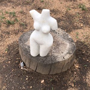 Artistic white sculpture of a female torso displayed on a wooden stump, surrounded by sandy soil and patches of grass in a natural outdoor setting.