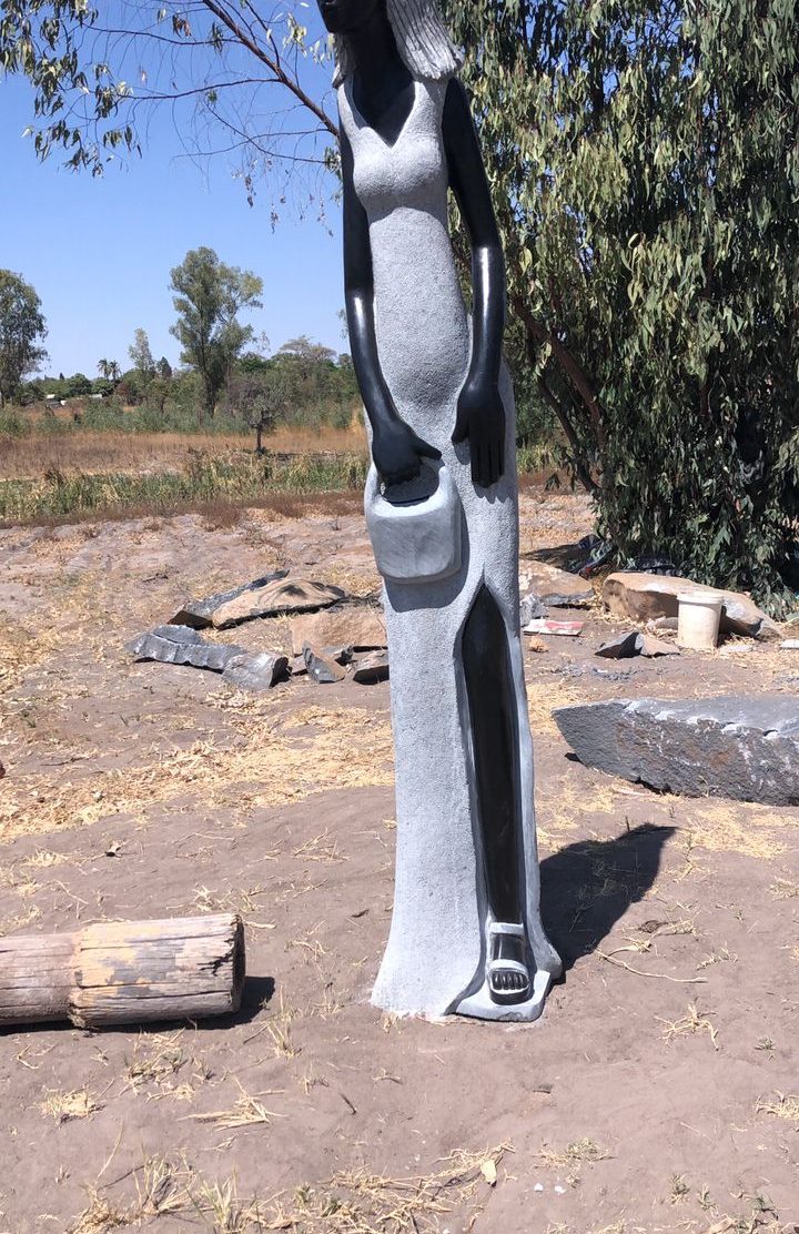 Sculpture of a graceful woman in a fitted dress holding a small container, set in a natural outdoor landscape with dry grass and stones.