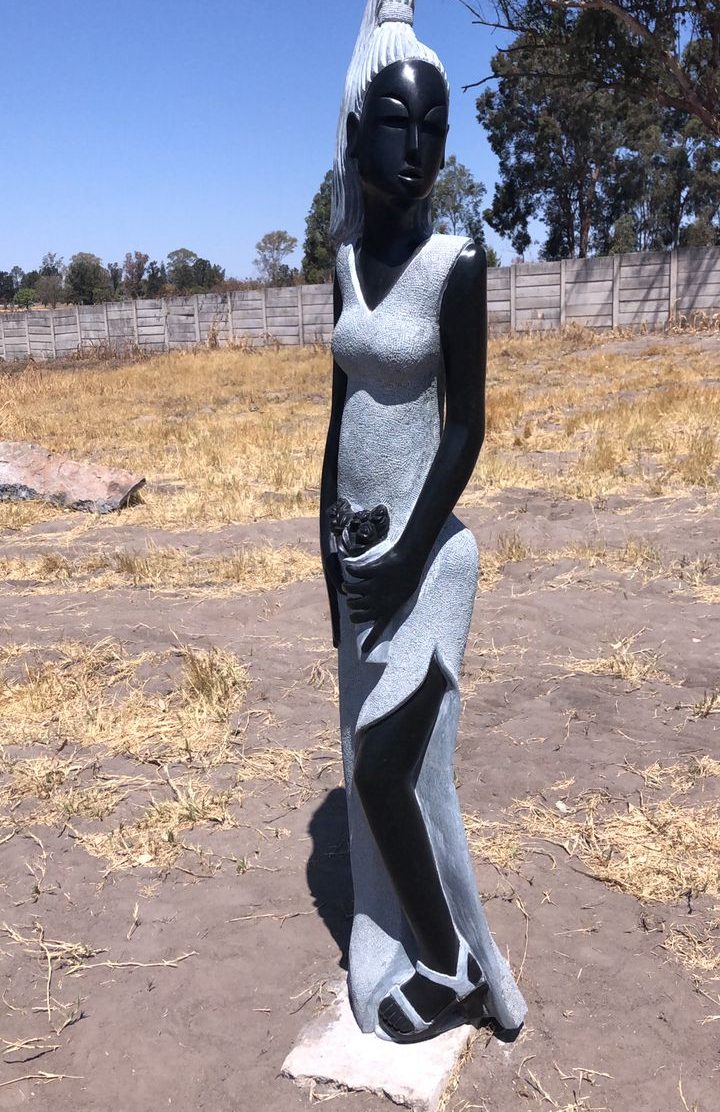 Tall black and grey sculpture of a female figure in a gown, set against a blue sky in a grassy outdoor environment