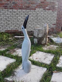 A white bird statue resembling a heron or crane stands amidst overgrown grass with brick walls in the background, perfect for garden decor and bird-themed art discussions.