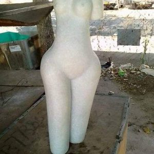 Light-colored humanoid mannequin standing on rugged surface, featuring a smooth texture and simplified human form without facial details, set against a blurred outdoor background.