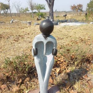 Unique abstract stone sculpture with heart-shaped torso and glossy head, surrounded by dry leaves and open grassy area under a clear blue sky.