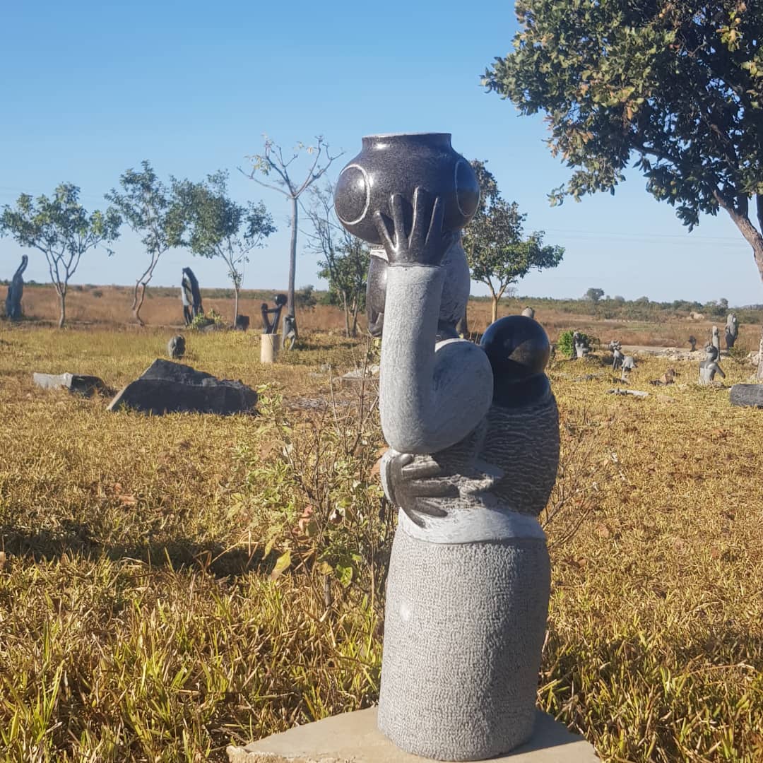 A detailed carved stone sculpture depicting a Mother holding a traditional clay pot (hari) on her head, and a baby on her back, set in a grassy area with sparse trees and other sculptures under a clear blue sky.