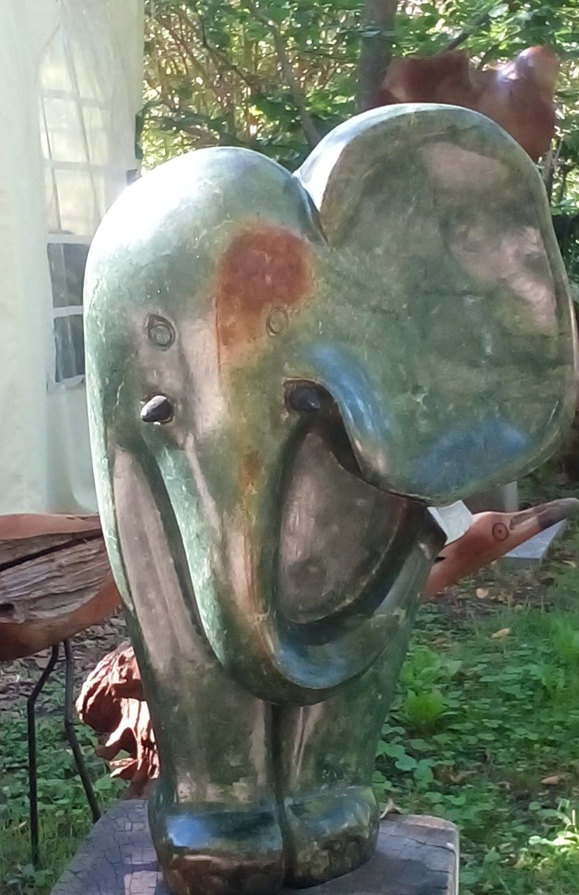 A beautifully carved elephant sculpture with a polished surface and a raised trunk, surrounded by a serene green wooded area.