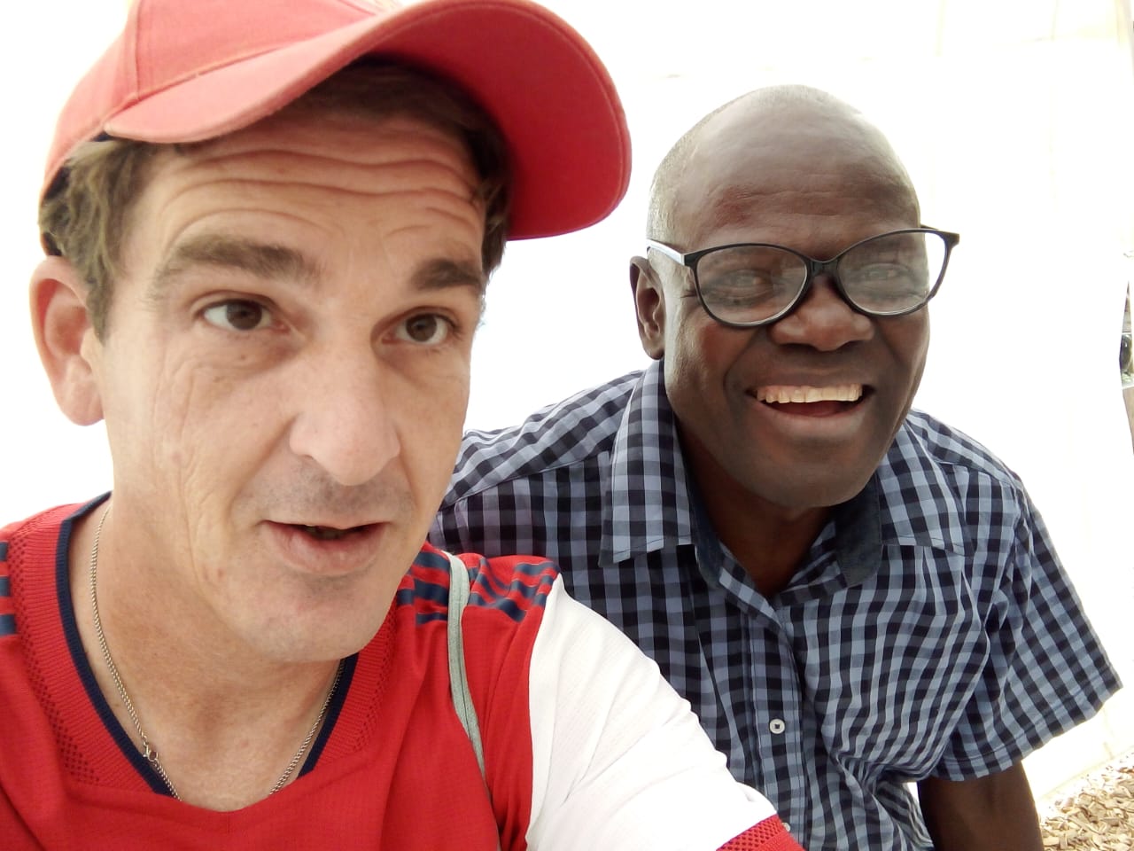 Two men smiling together outdoors, one in a red cap and shirt, and the other in glasses with a checkered shirt, on a blurred background.