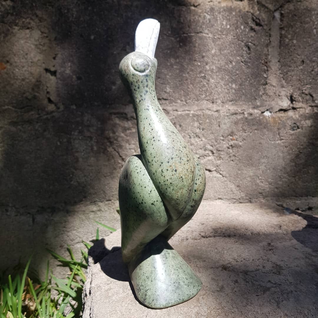 Stylized green stone sculpture of an African Fish Eagle - Hungwe - the Zimbabwean National bird with a white beak, placed on a stone surface, illuminated by natural light against a textured wall and grass.