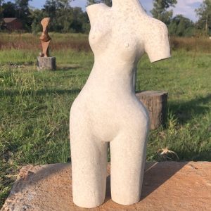 Artistic sculpture resembling a minimalist human torso made from light-colored stone, featuring a bird-like element, placed on a rustic wooden platform in a grassy outdoor area with abstract sculptures in the background.