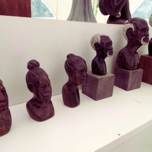 Collection of handcrafted wooden sculptures depicting human busts with various hairstyles and facial expressions, displayed on a white shelf under soft natural light.