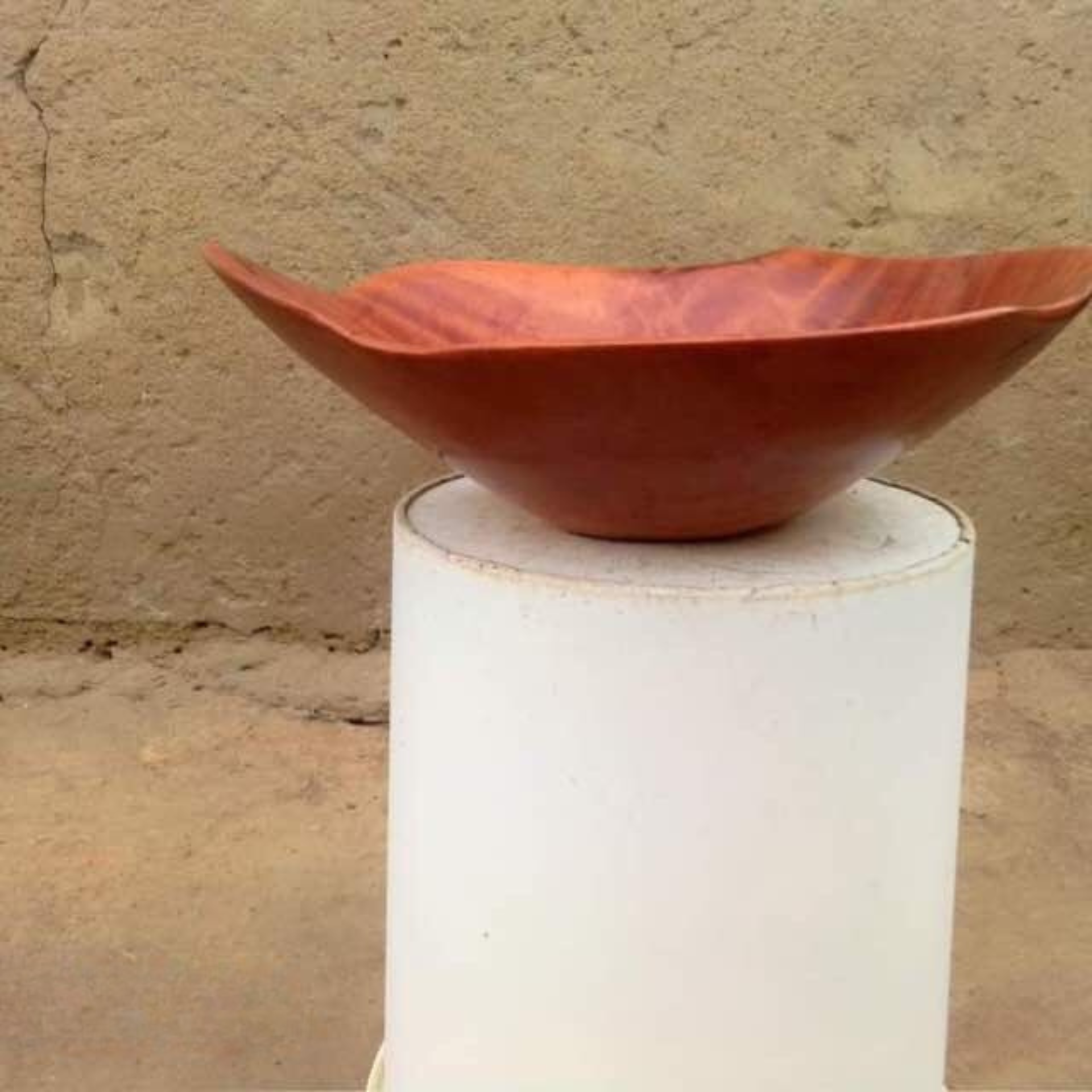 Elegant handcrafted wooden bowl with a wavy edge and rich grain displayed on a white pedestal against a textured beige background.
