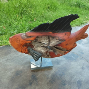 Artistic wooden sculpture of a fish with polished surfaces and detailed grain patterns, displayed on a metal base against a grassy outdoor background.