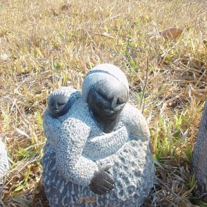 A stone sculpture of a kneeling woman in a grassy area, wearing traditional dress, with a small child resting on her back; surrounded by dry grass and distant stone figures.