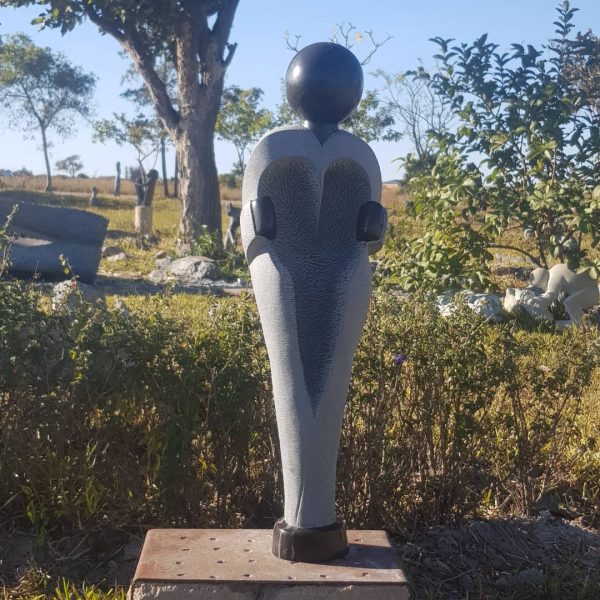 A striking gray sculpture with a heart shape at its center and a black spherical head, surrounded by grass and shrubs under a clear blue sky.