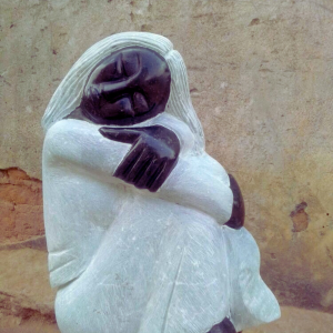 Close-up of a beautifully carved sculpture depicting a seated figure with a serene expression, made of dark stone and translucent light stone, hugging its knees against a textured wall.