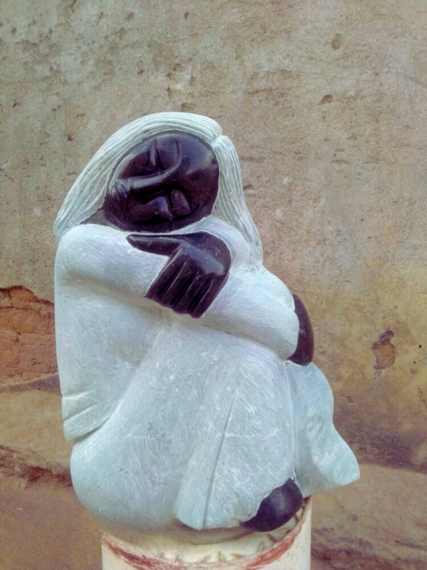 Close-up of a beautifully carved sculpture depicting a seated figure with a serene expression, made of dark stone and translucent light stone, hugging its knees against a textured wall.