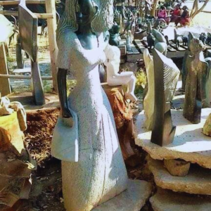 A stunning outdoor display of various sculptures including a prominent stone sculpture of a woman wearing a flowing dress, surrounded by a natural setting with trees and an earthy ground.