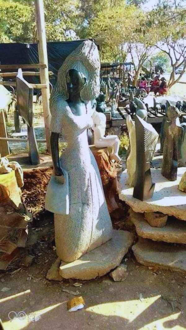 A stunning outdoor display of various sculptures including a prominent stone sculpture of a woman wearing a flowing dress, surrounded by a natural setting with trees and an earthy ground.