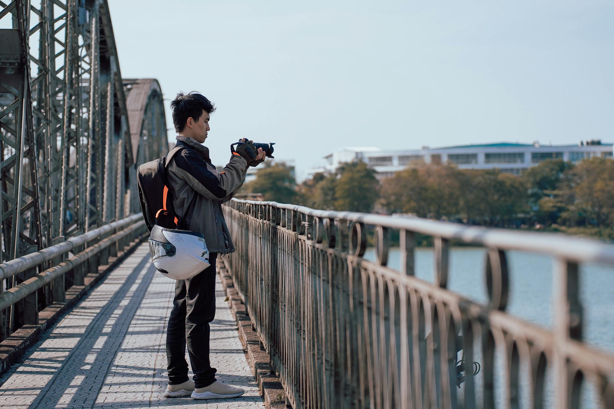 Asian man capturing scenic views on a metal bridge with a camera, during a sunny day.